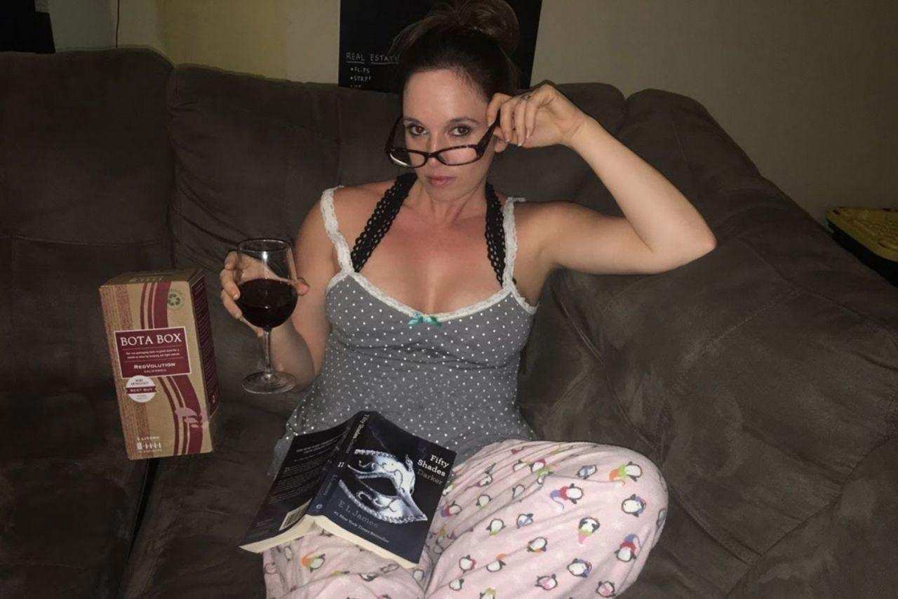 A woman sitting on the couch in her pajamas with a glass of wine and 50 shades of gray book on her lap